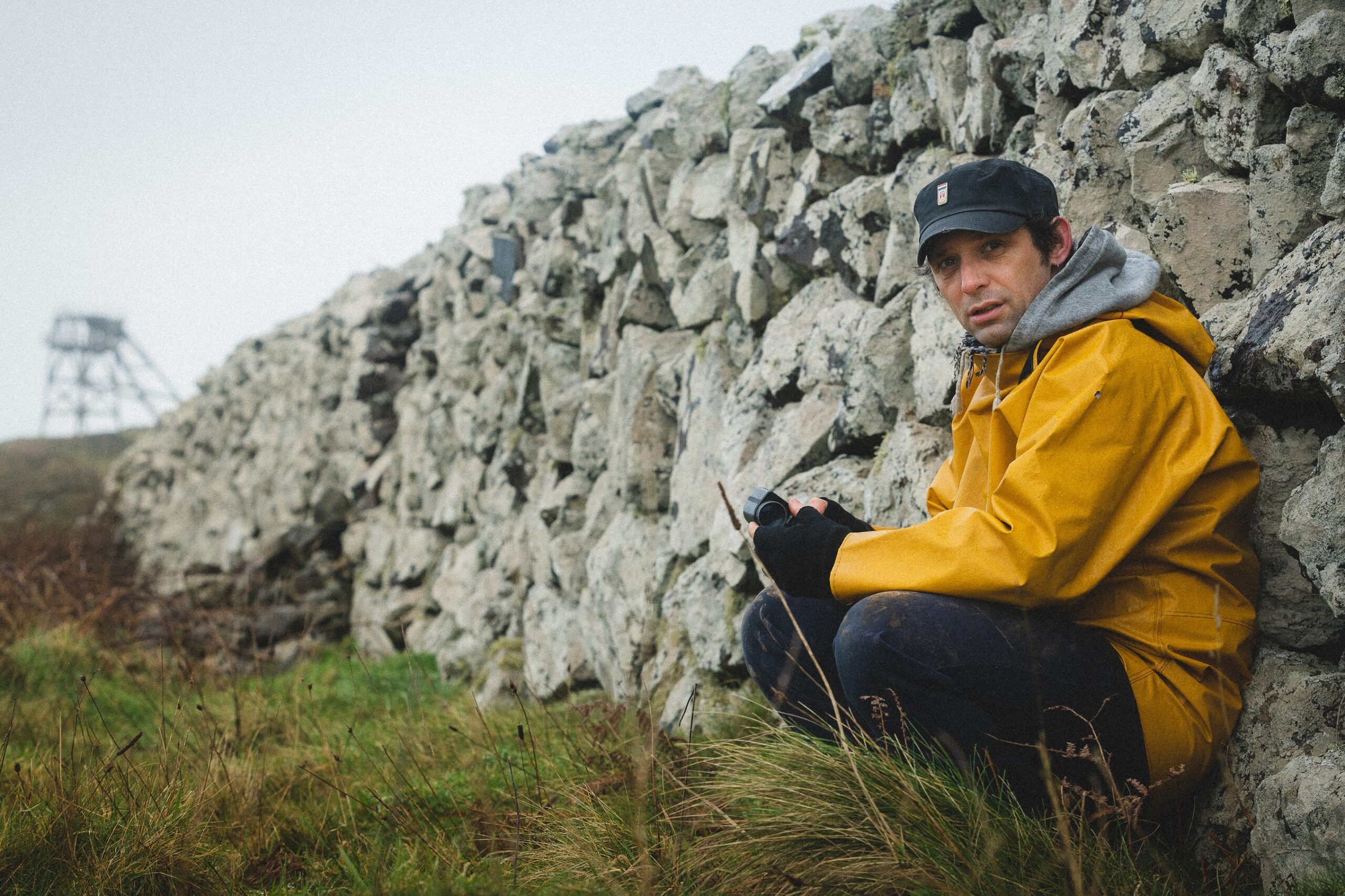 Mark Jenkin in a yellow rain coat sits against a stone wall